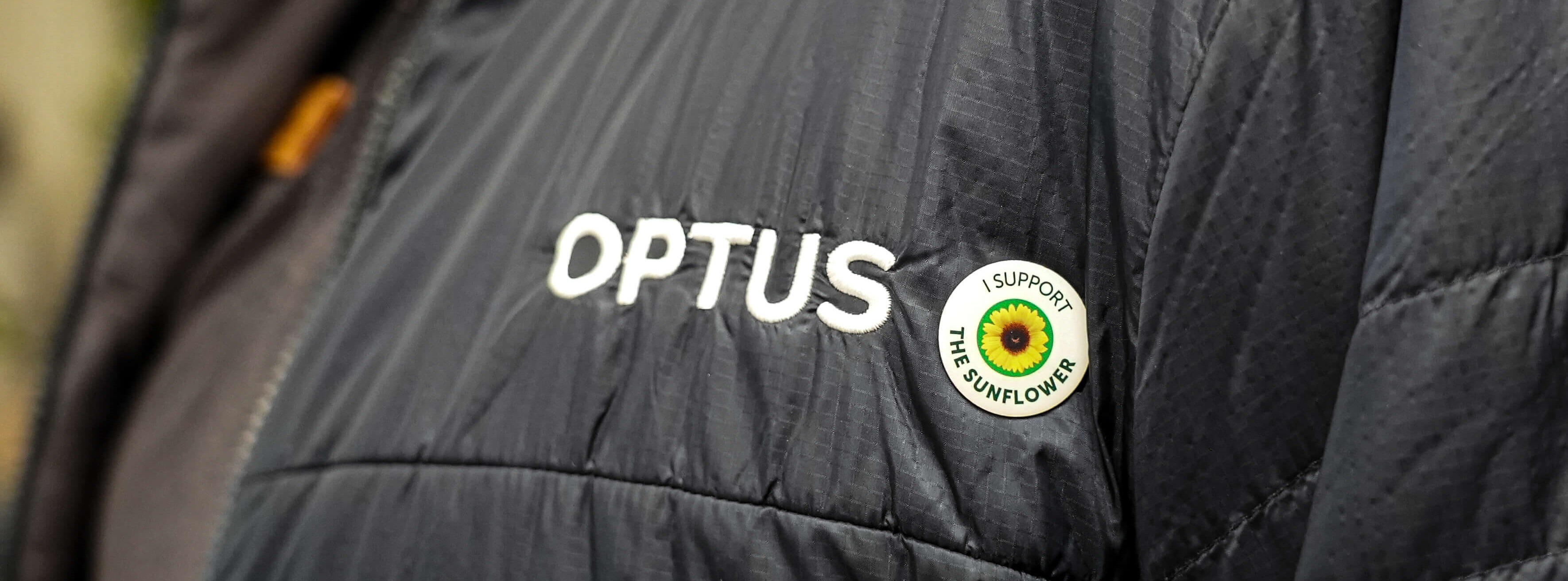 Extreme close up of an Optus employee's jacket. Next to the Optus logo on the jacket, the employee has pinned an 'I Support The Sunflower' badge.
