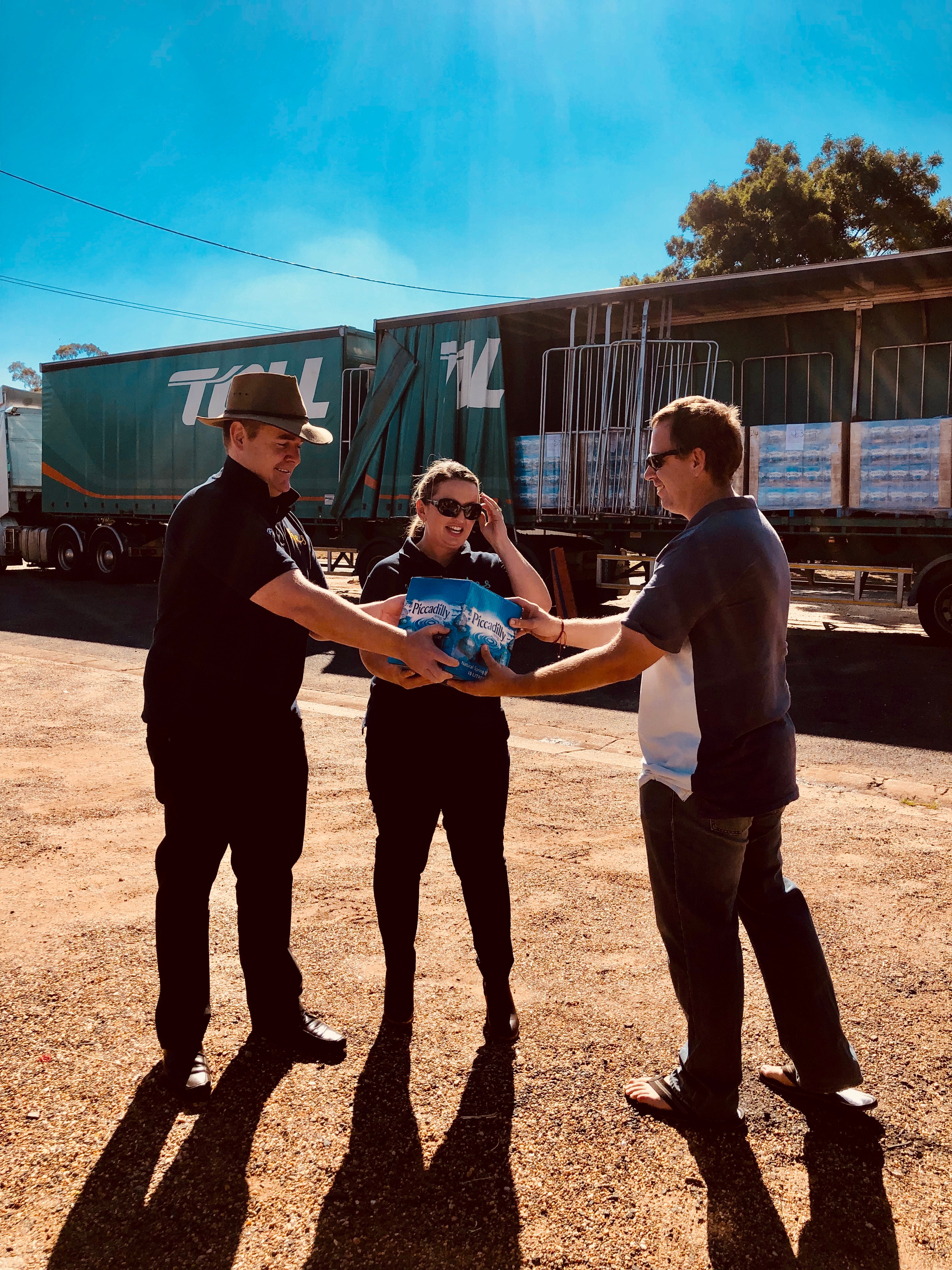 A man and a woman hand a box of water to another man. In the background is a Toll truck with pallets of water boxes. 