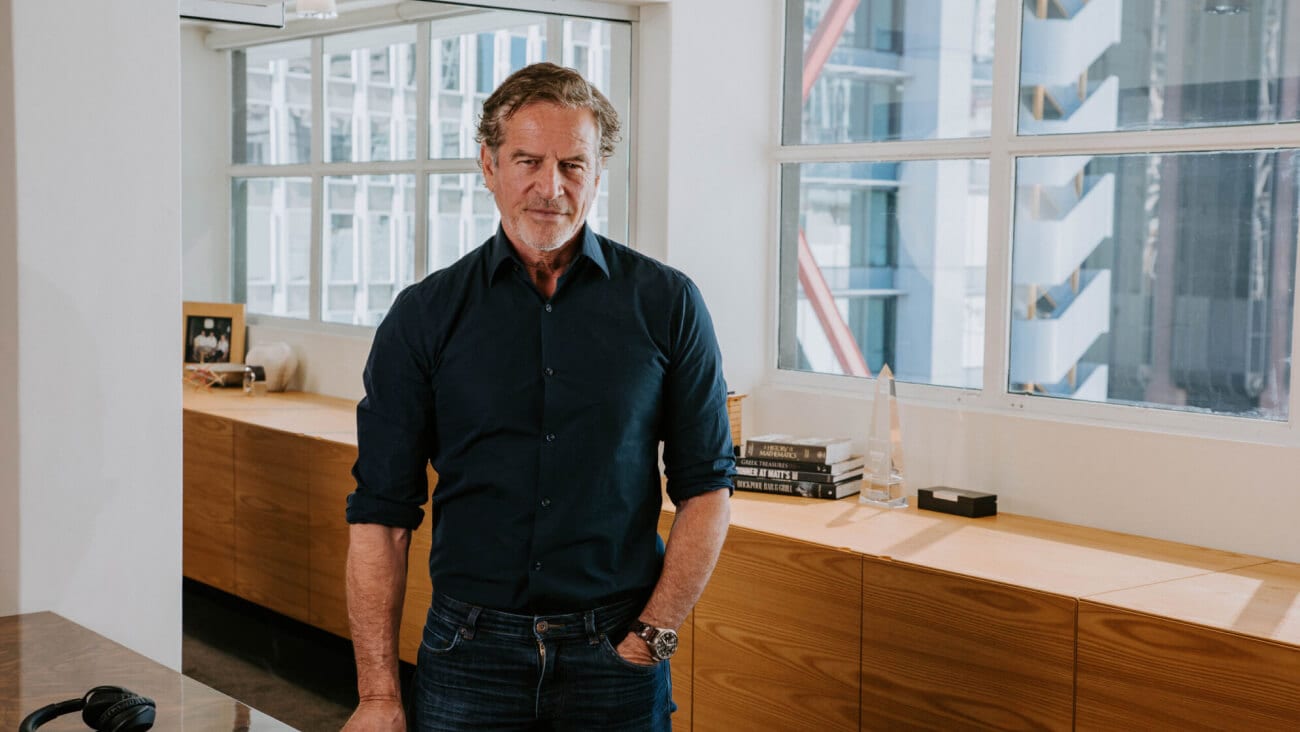 Mark Bouris in a dark shirt and trousers standing in an office with light coming in from the windows.