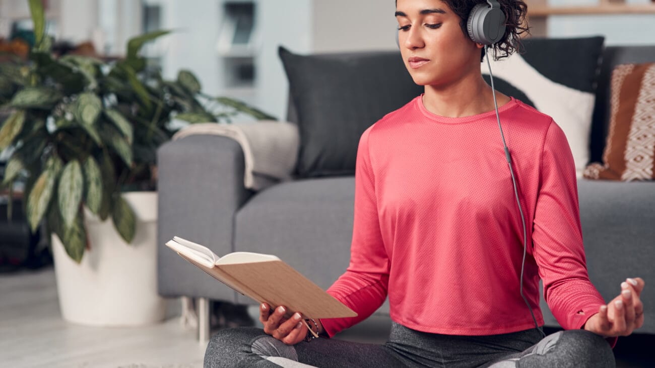 Young woman with headphones sitting in a yoga pose on the floor reading a book. 