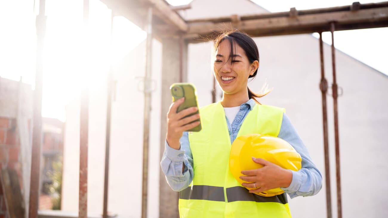 Young female tradesperson in a high visibility vest holding a mobile phone while onsite.
