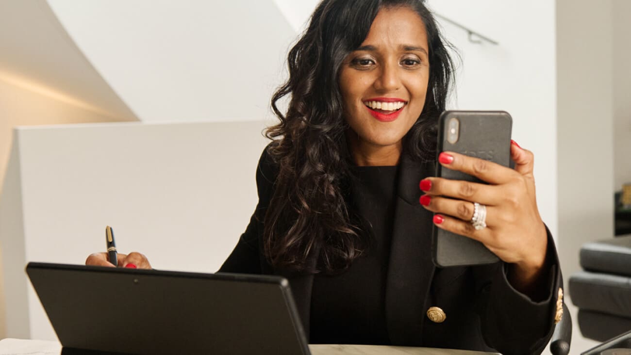 Young woman with long dark hair in a business attire sitting at a desk with laptop using a mobile. 