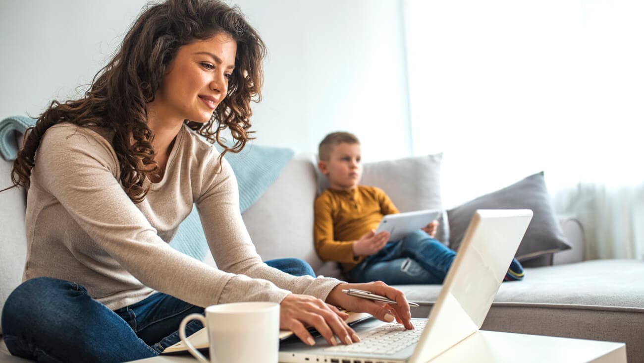 Mother on a laptop sitting on a sofa with son in the background using a tablet.