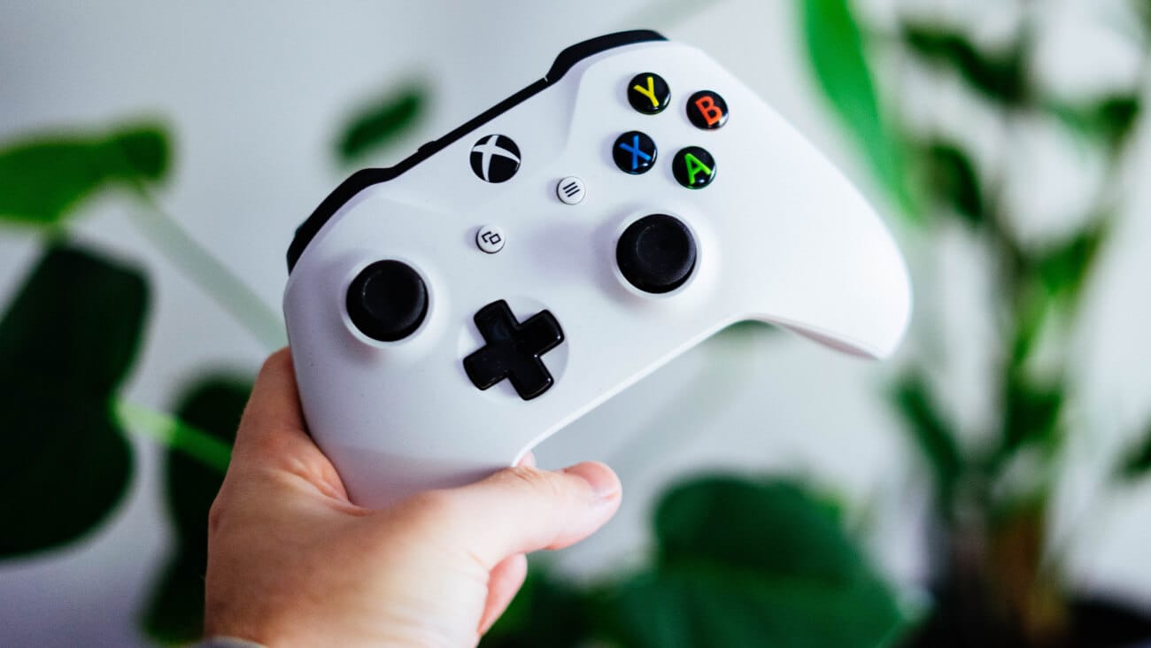 White wireless Xbox controller held diagonally by a hand with a blurred background of plants with green leaves.