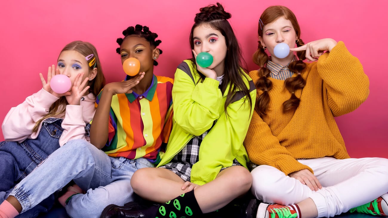 Four teen girls blowing colourful gum bubbles with a pink background.