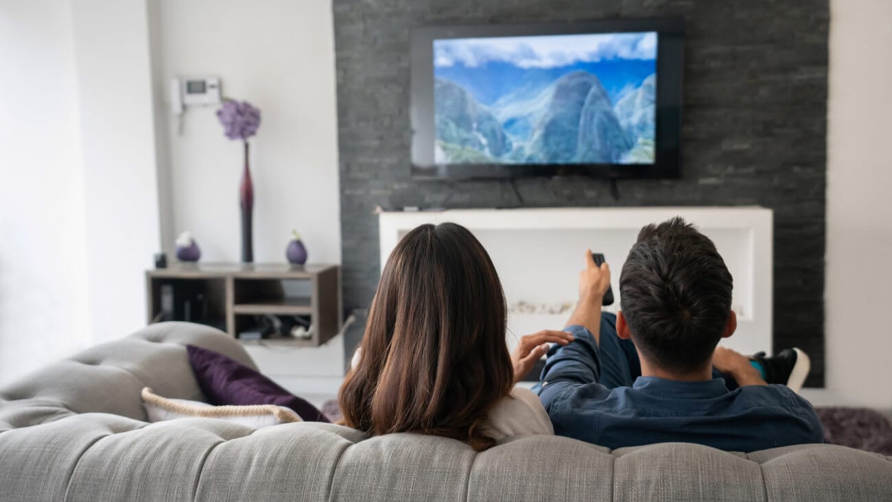 Couple facing home cinema screen showing a mountainous scene, sitting on a light grey lounge.