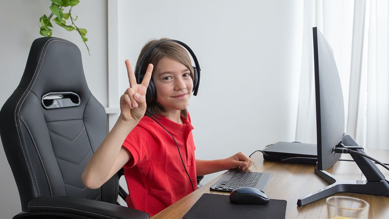 Young boy in red shirt sitting on black chair at a desk with a computer monitor.