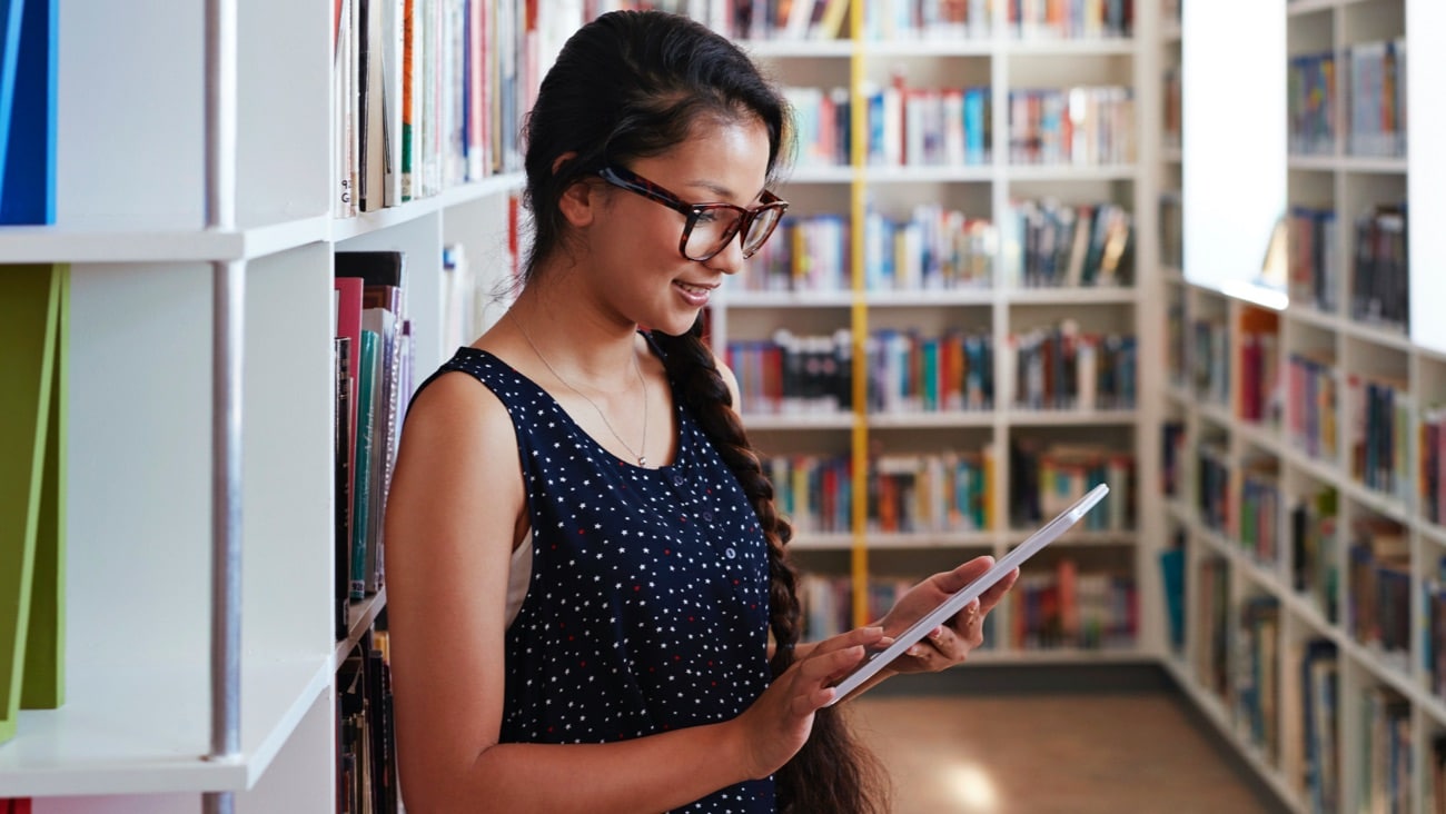 Girl looking at tablet with books behind her.