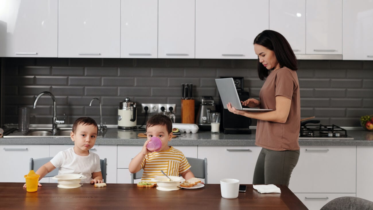 A woman using her laptop while walking near her kids eating a meal at the kitchen table