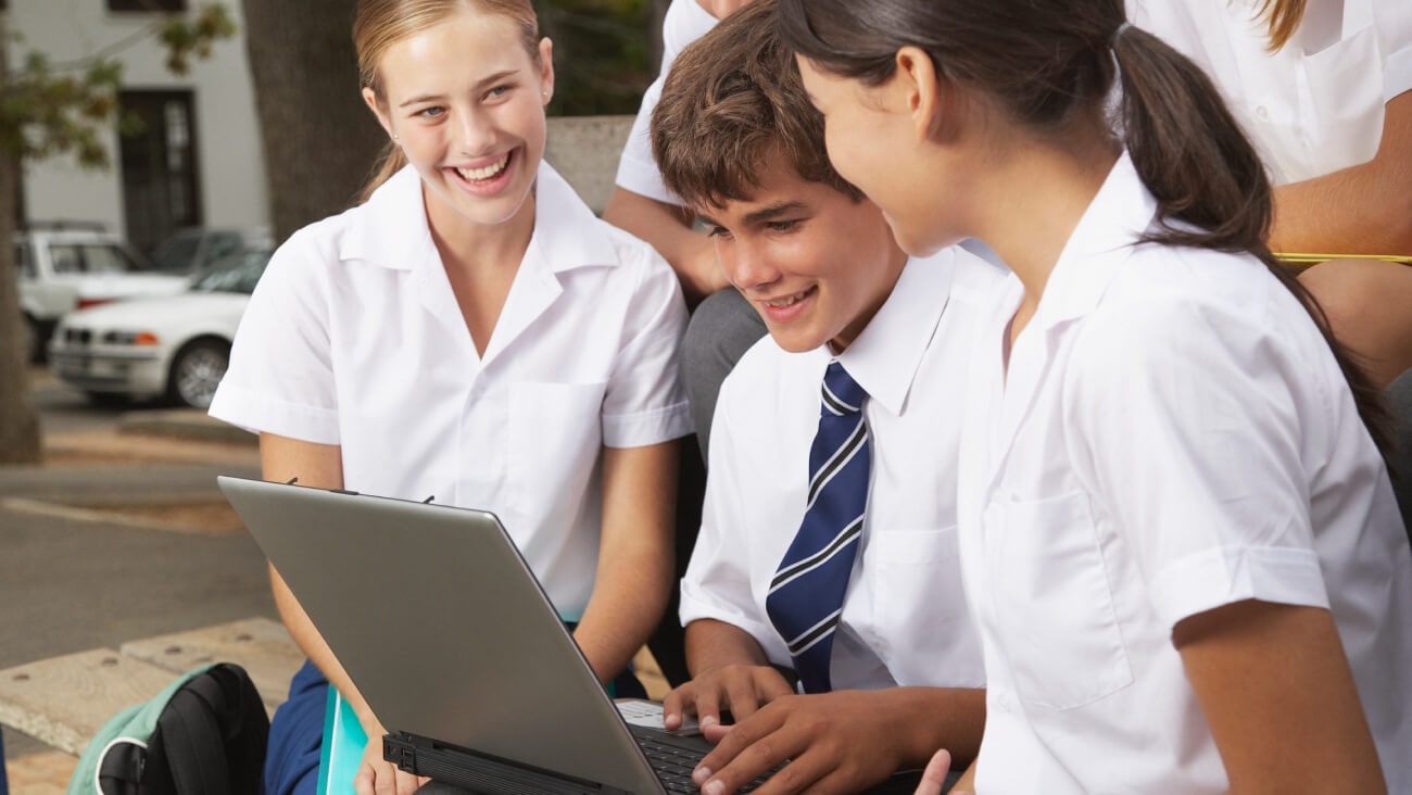 Two girls and a boy working on a laptop outside with other students around them.