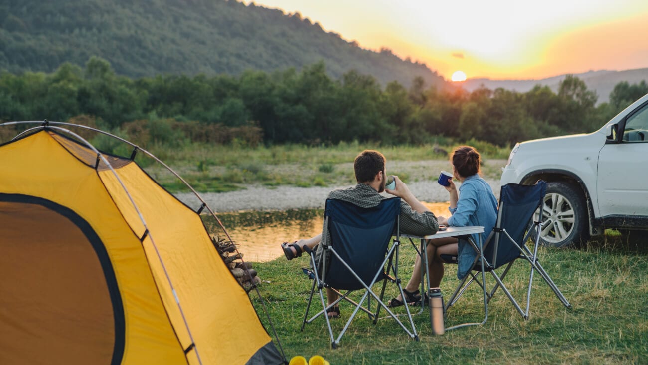 Couple sitting in camp chairs looking at sunset above river in mountains.