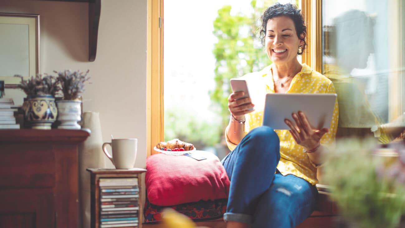 Woman is sitting by a window smiling and holding a phone and tablet