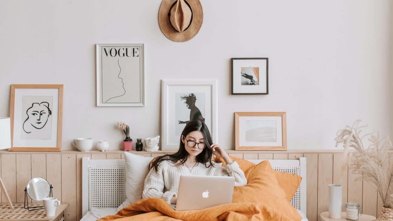 young woman on her laptop sitting in a bed surrounded by prints on the wall