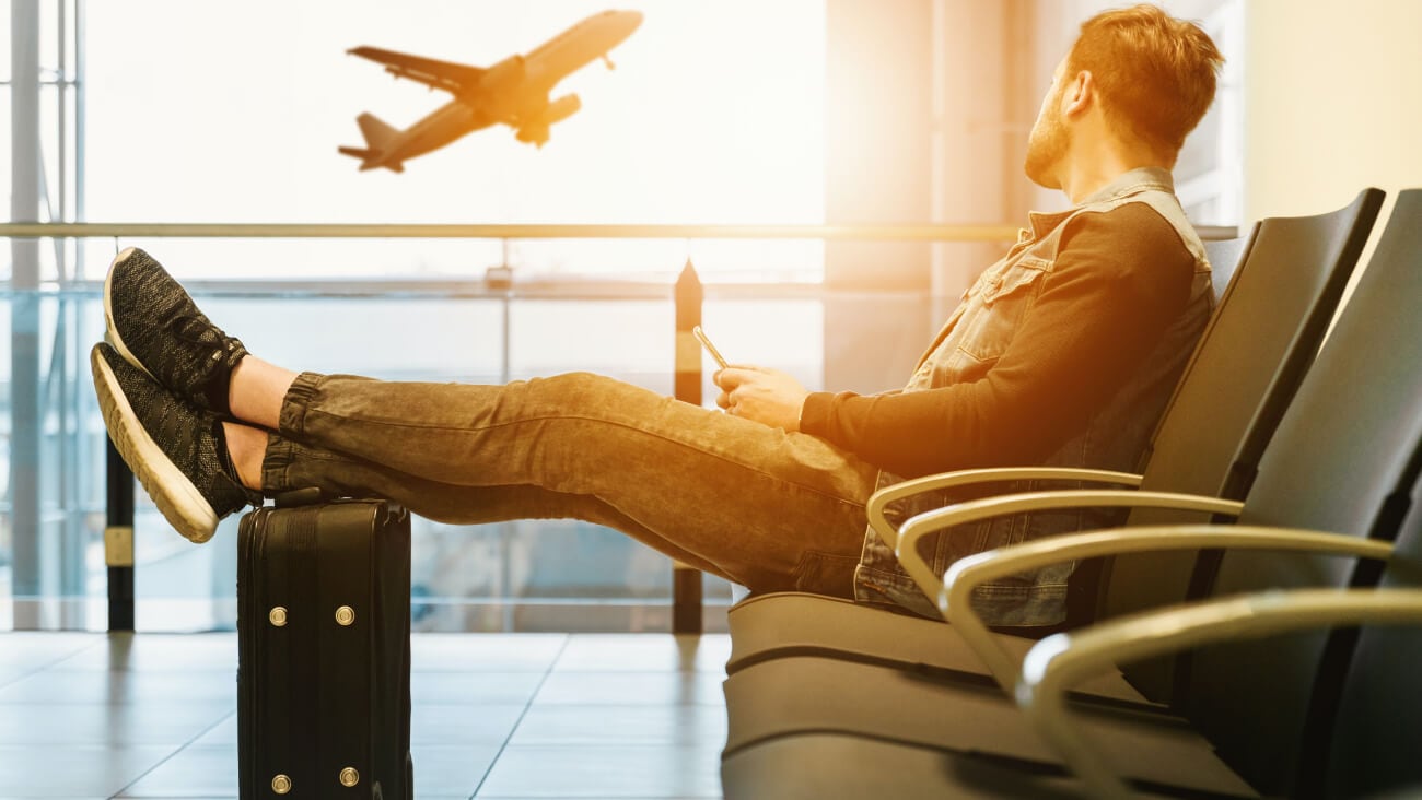 man sitting at airport with feet up on luggage, looking out window at plane taking off