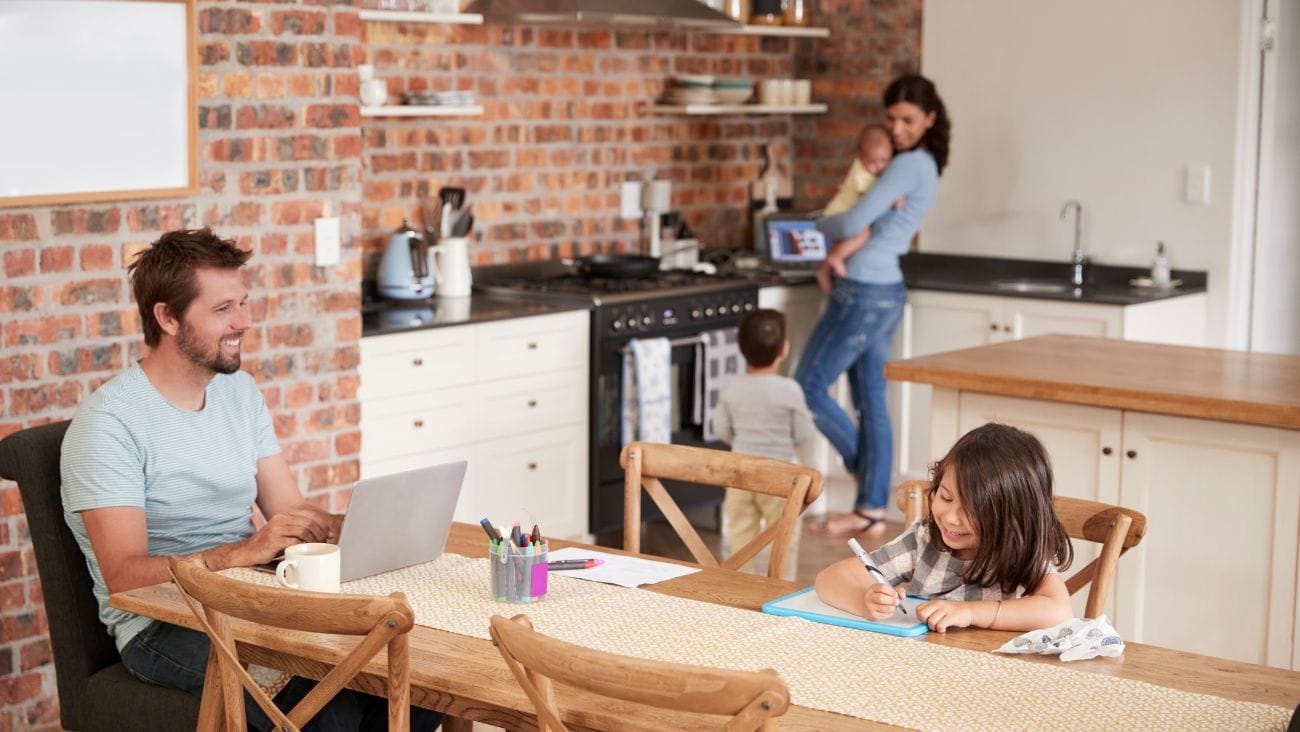 Father sitting at the dining table with a laptop and his daughter drawing while his wife stands in the kitchen with their baby.