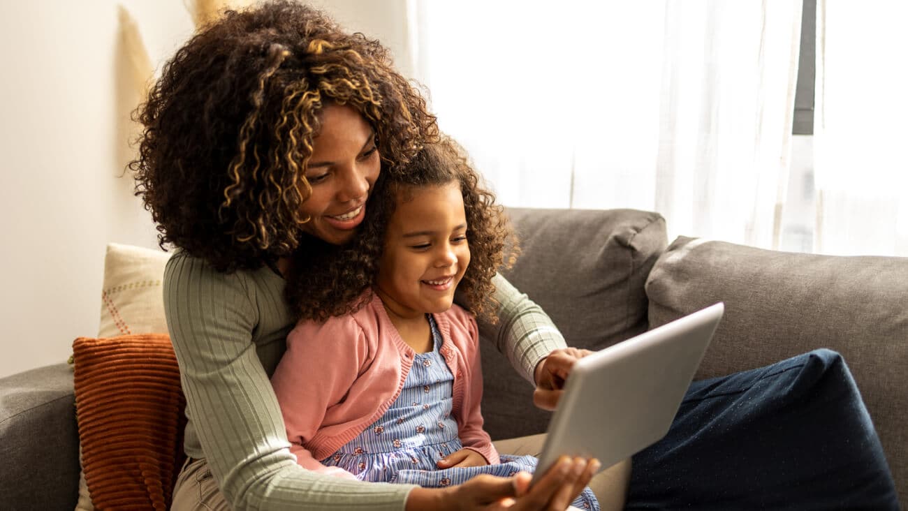 Mum and daughter sitting on a couch smiling while using a tablet