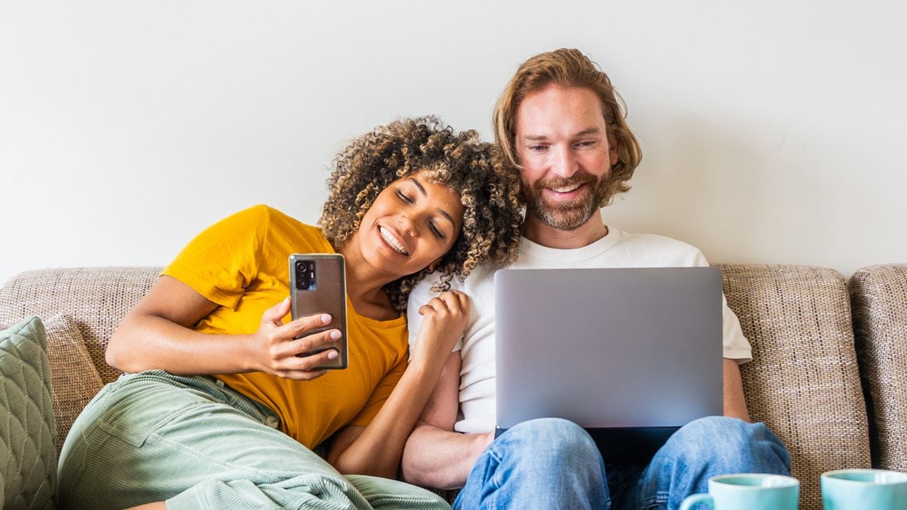 A man and woman smiling and looking at a phone and laptop while sitting on a couch.