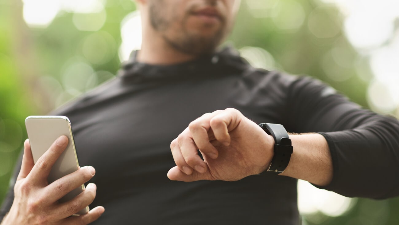 man in black activewear looks at watch on left wrist while holding a grey smartphone in his left hand