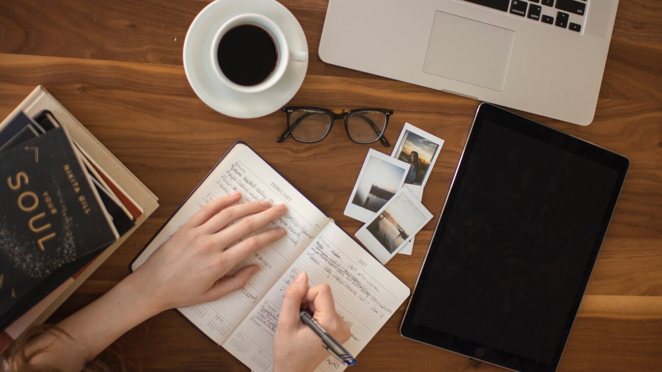 laptop and stationery items on a table with a person writing in a diary