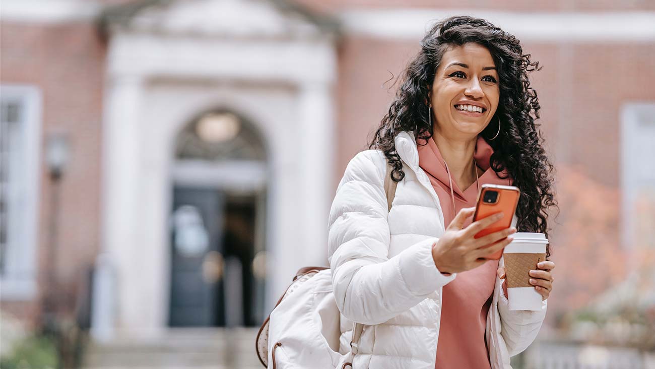 Young woman with long dark hair outside a university holding a phone and a takeaway coffee
