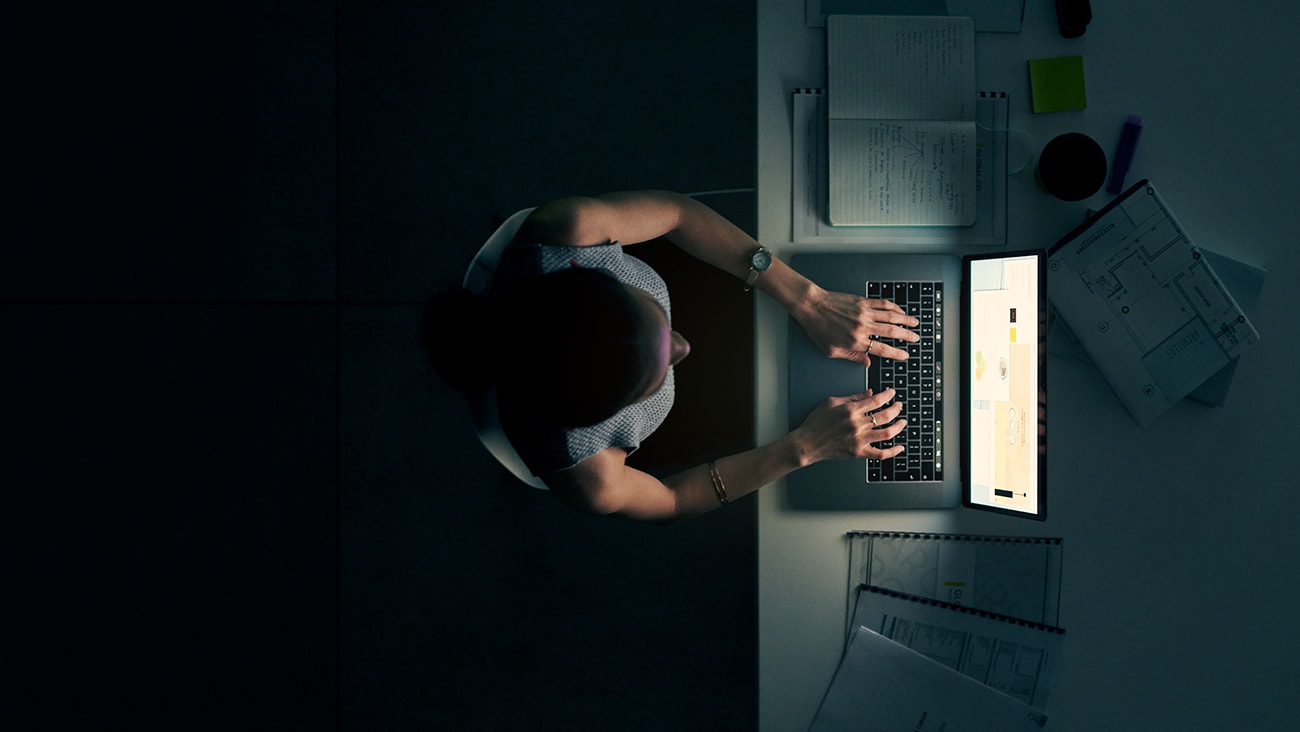 A woman working at a laptop in dark conditions, the laptop screen is the only light-source in the room
