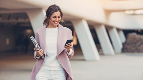 Young woman walking outside an office building wearing a pink coat using a mobile phone.