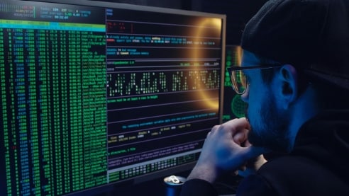 An online hacker sitting in the dark in front of computer screens.