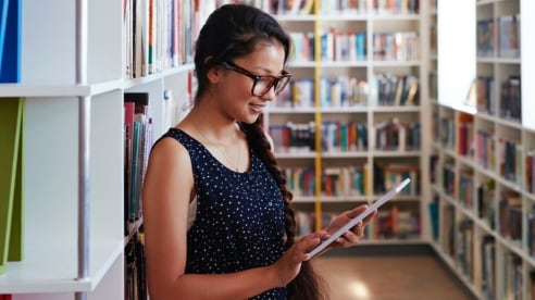 Girl looking at tablet with books behind her.