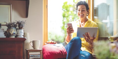 Woman is sitting by a window smiling and holding a phone and tablet